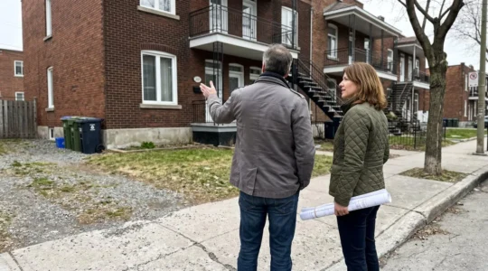 Couple dans la quarantaine debout devant une maison en brique rouge typique de Montréal, discutant d'un projet d'agrandissement en pointant vers le côté de la résidence